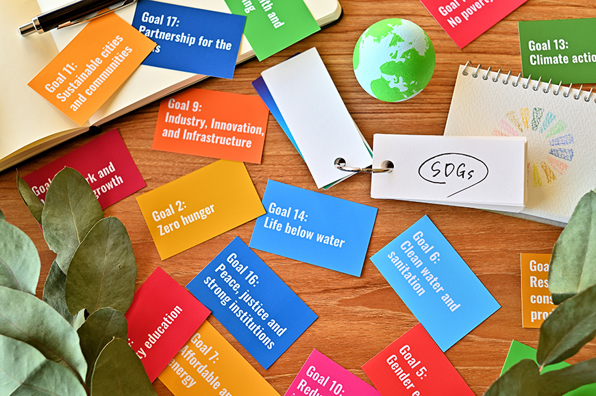 colorful cards displaying the united nations sustainable development goals spread across a wooden desk, alongside a small globe and a notepad labelled “sdgs