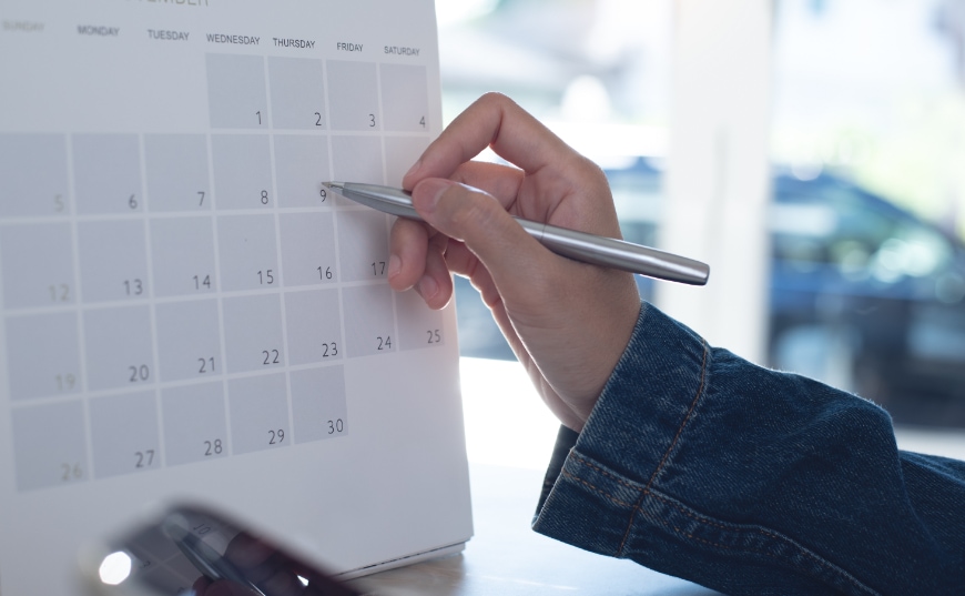 a hand holding a pen up to mark a date on a wall calendar.