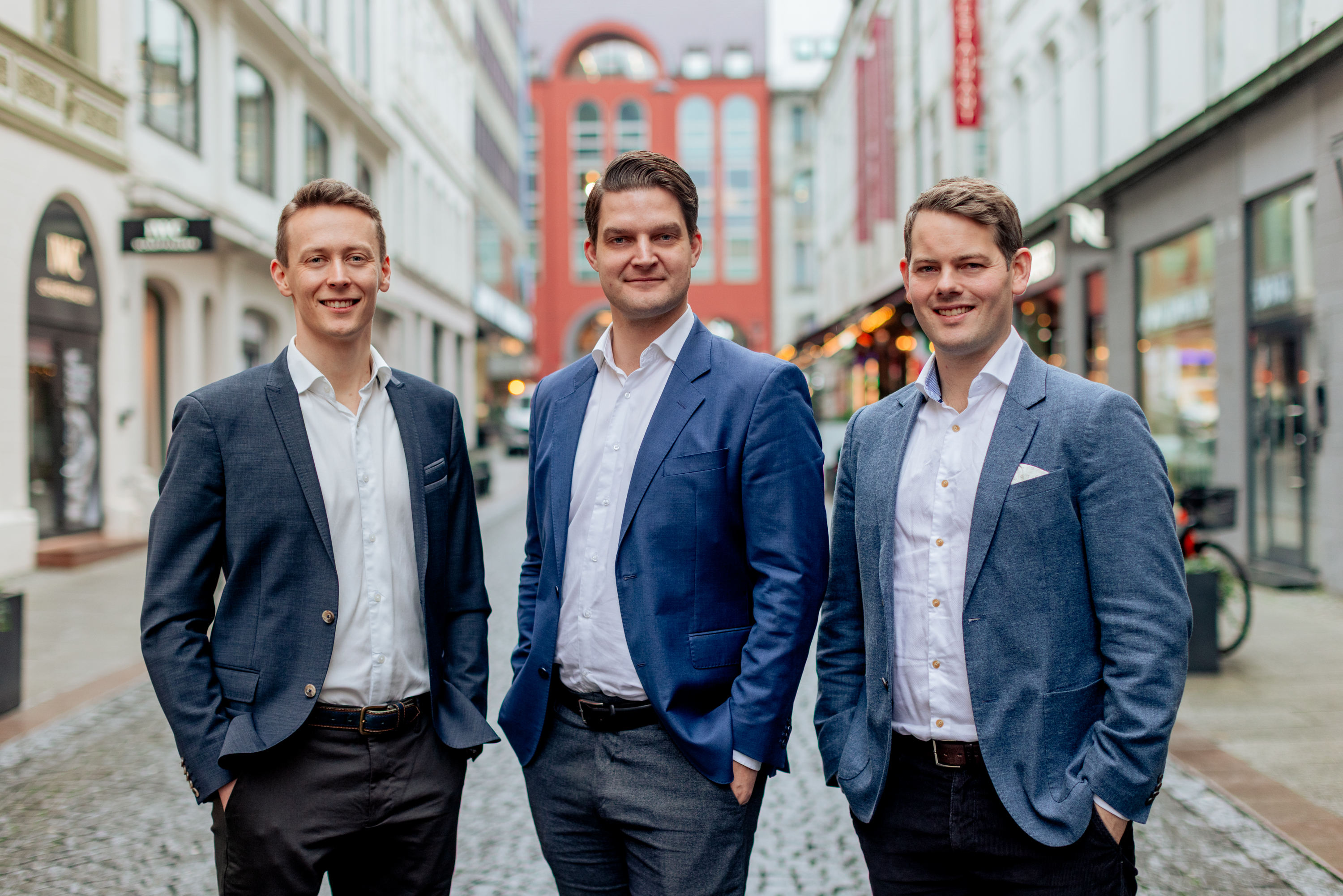 Three men in business attire standing on a cobblestone street with buildings and shops in the background.