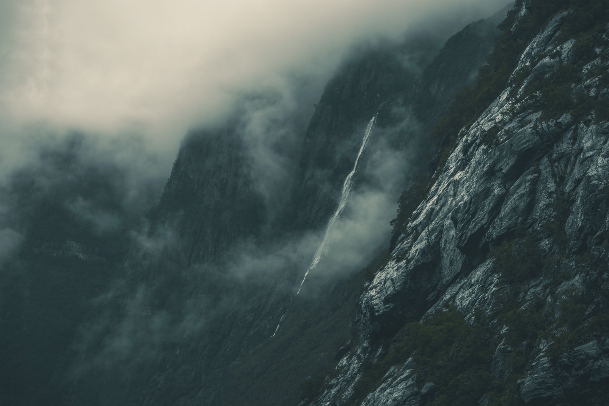 Misty mountain slopes with a narrow waterfall cascading down rocky cliffs.