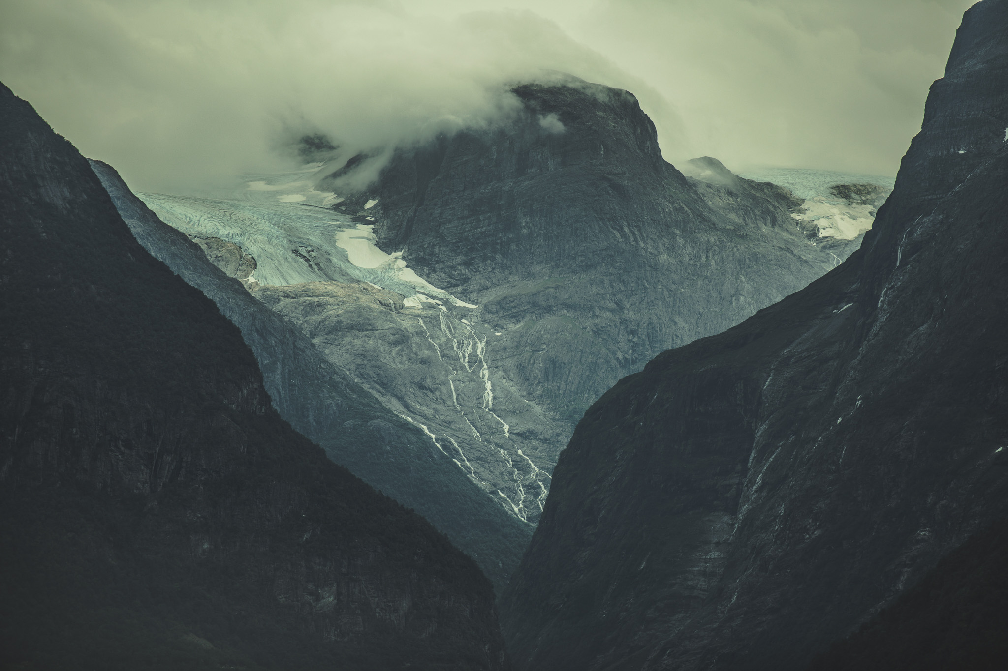 Fog-covered mountain peaks with a glacier flowing through a valley and streams of melting snow.