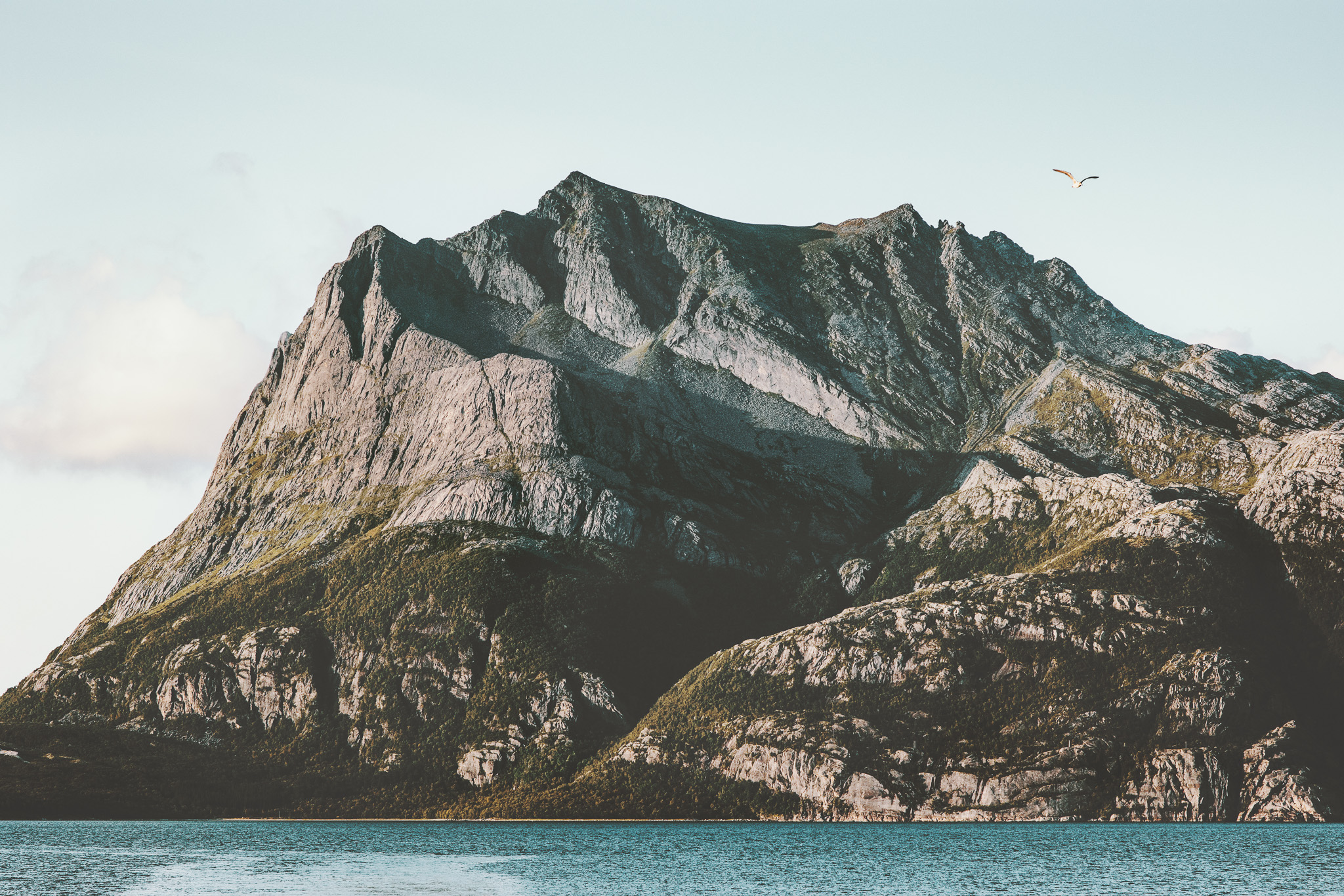 Large rocky mountain with rugged cliffs next to a calm blue body of water and a clear sky with a bird flying.
