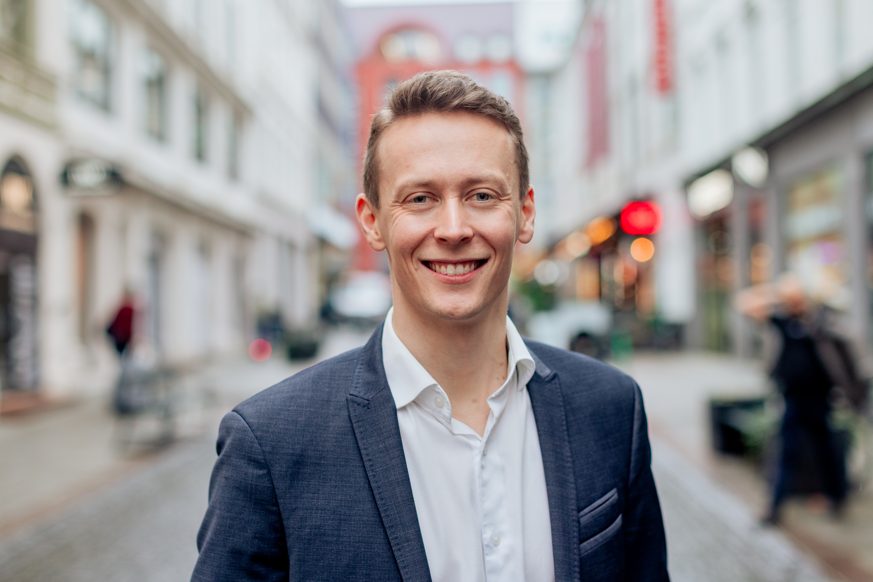 Smiling young man in a navy blue suit jacket and white shirt standing on a blurred city street.
