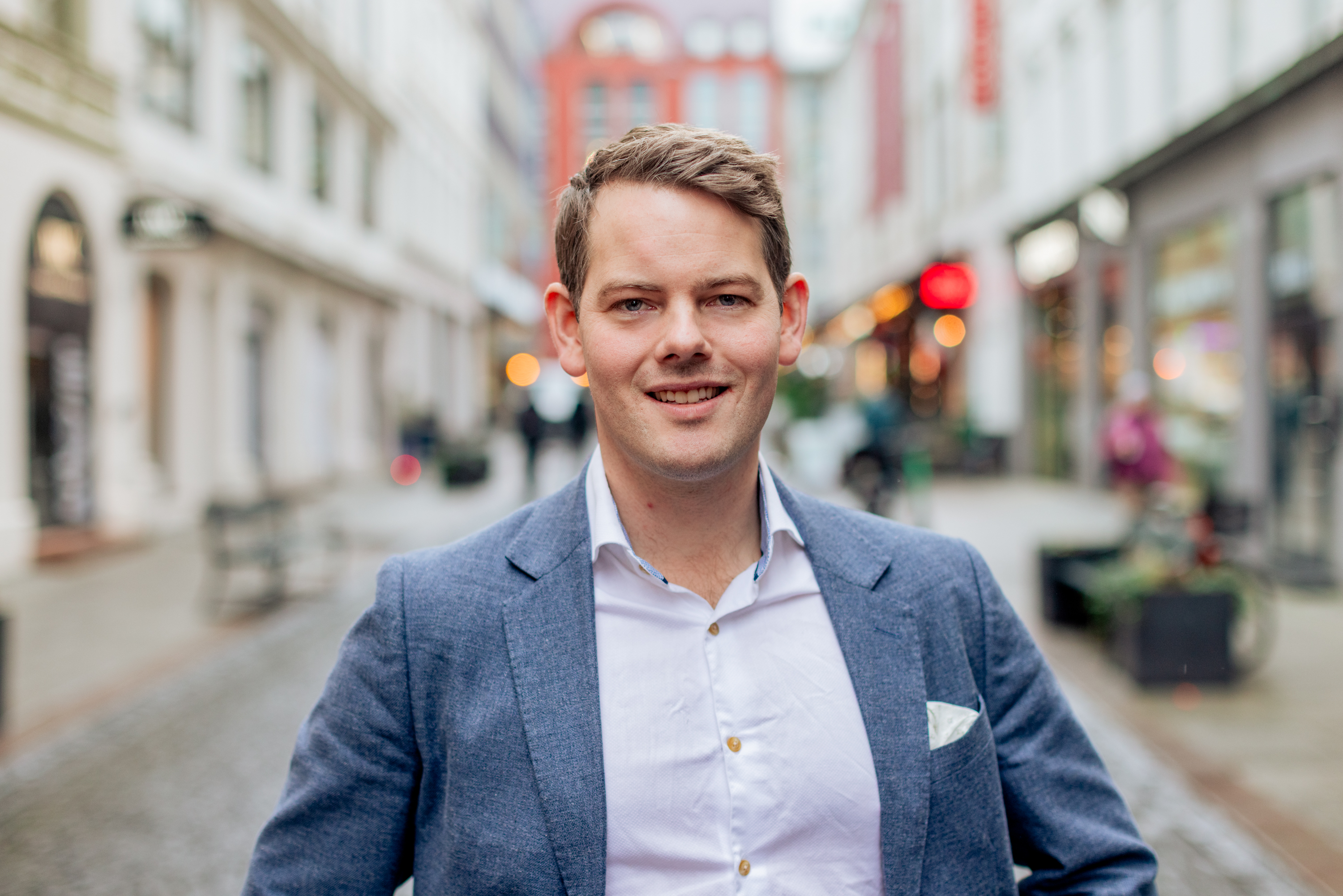 Smiling man in a blue blazer and white shirt standing on a city street with blurred buildings in the background.