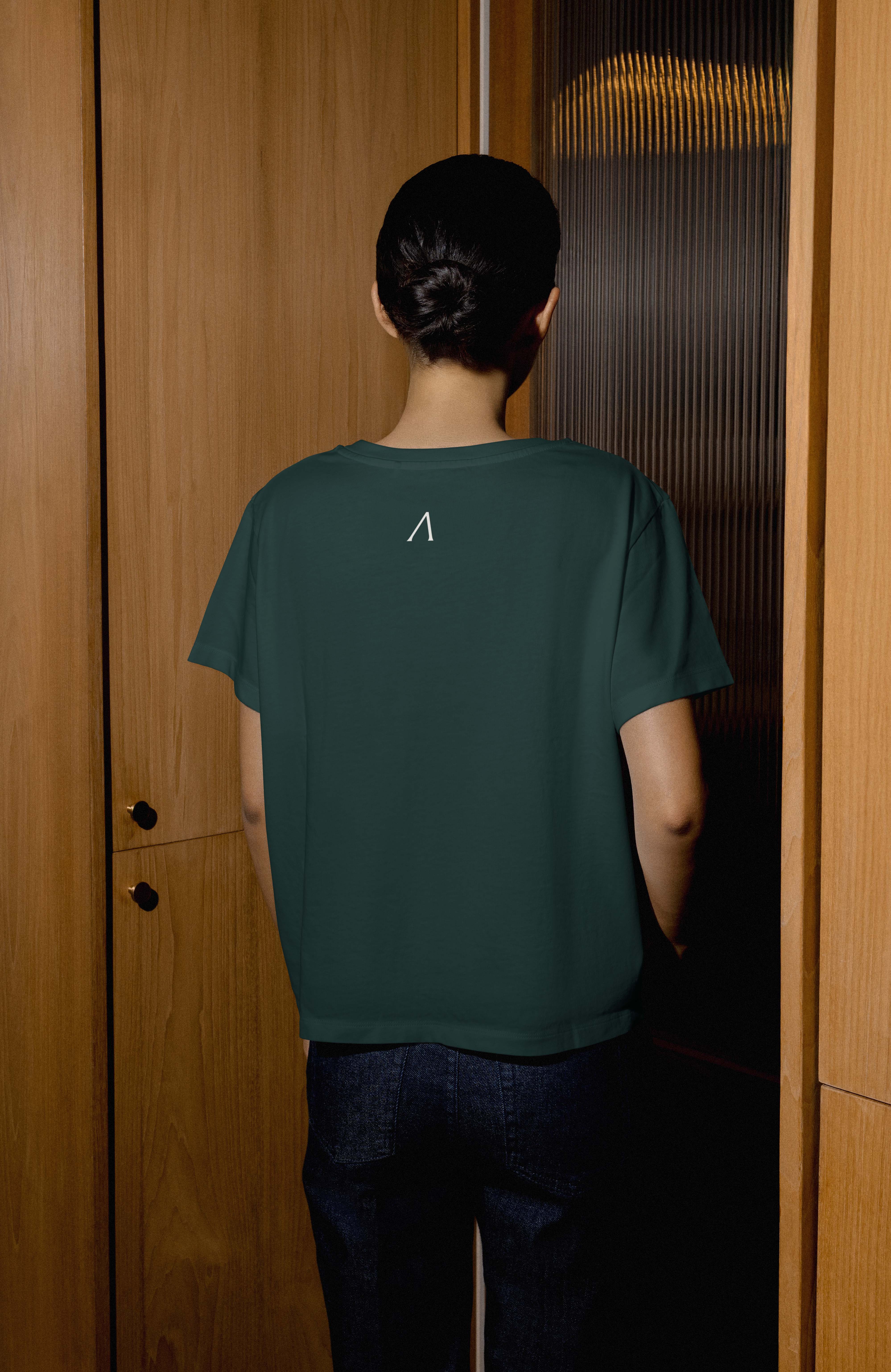 Person with dark hair in a bun wearing a dark green t-shirt with a small white symbol on the upper back, standing in front of wooden paneled walls.