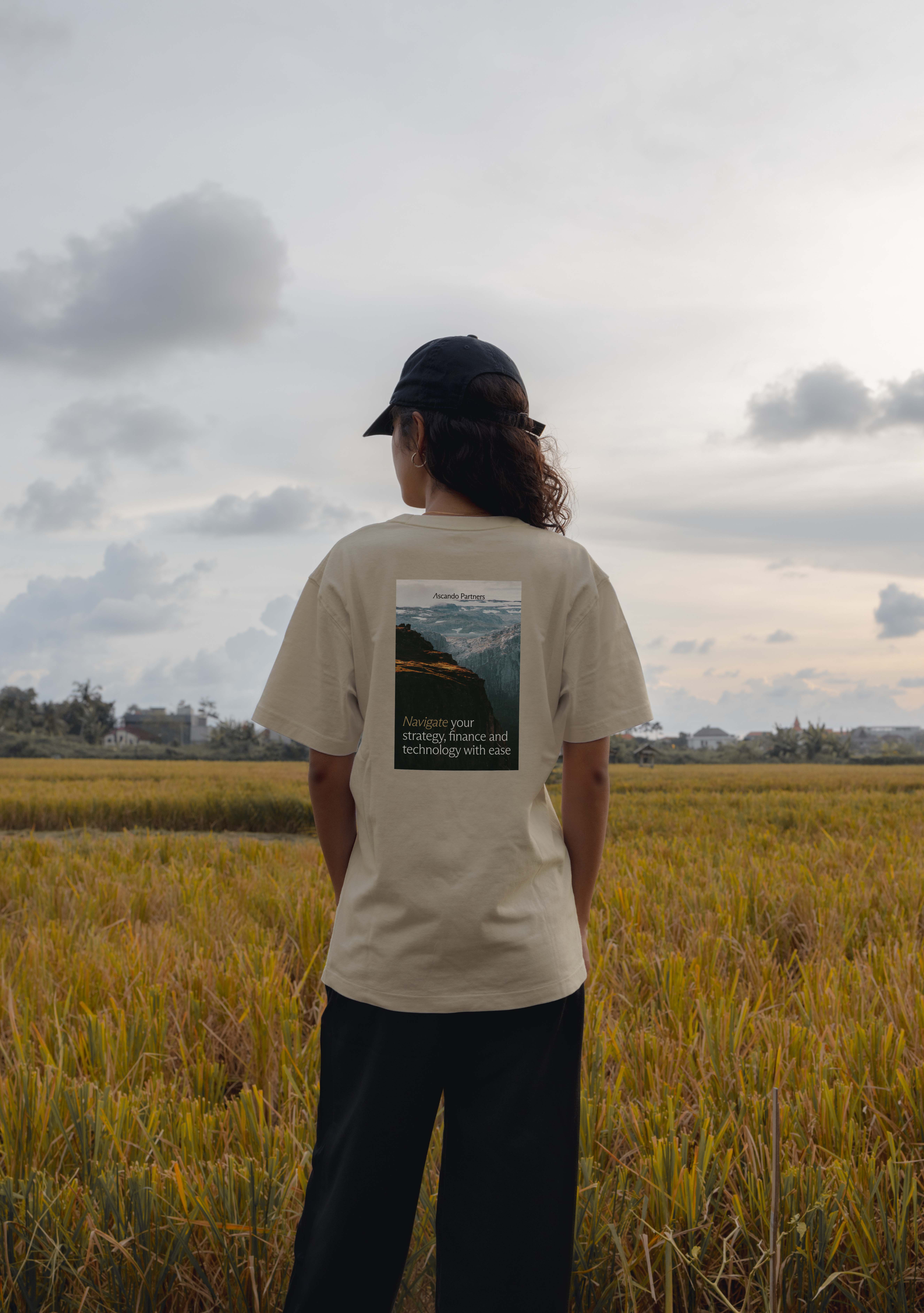 Person wearing a beige T-shirt with a mountain landscape and text on the back, standing in a field under a cloudy sky.