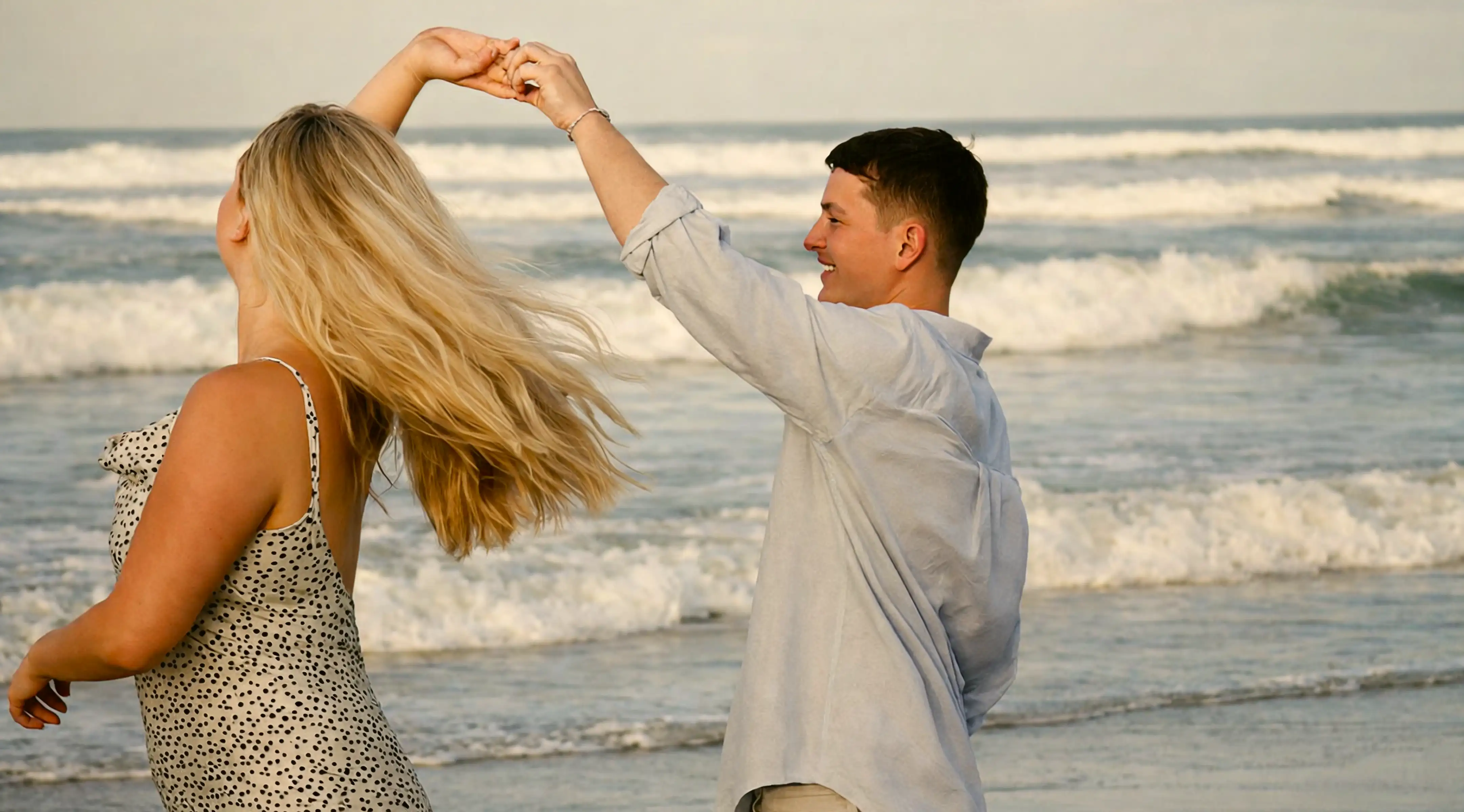 Couple dancing on a beach at sunset, hands raised as she spins