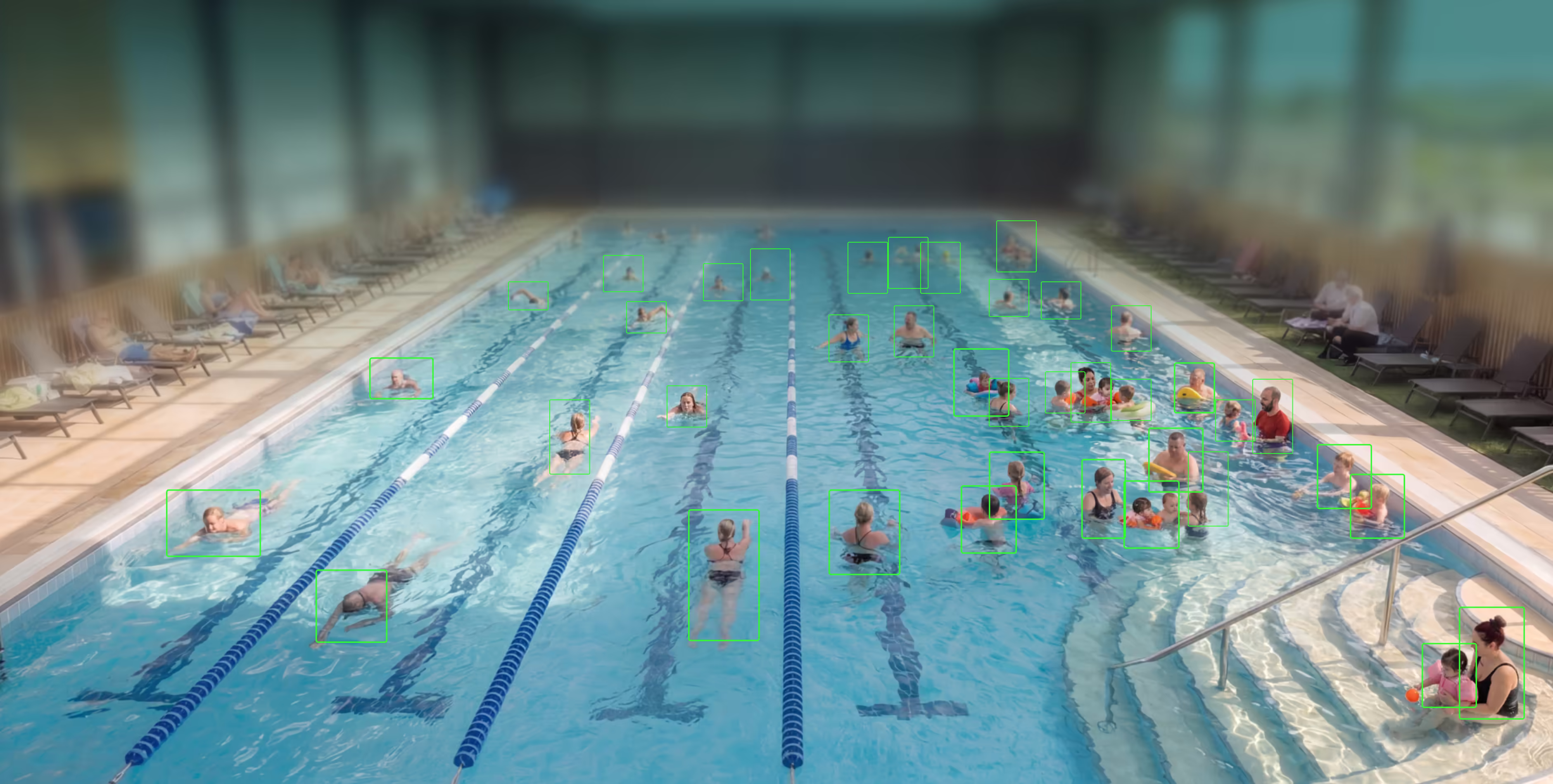 People of various ages enjoying swimming and playing in an indoor pool with onlookers seated nearby.