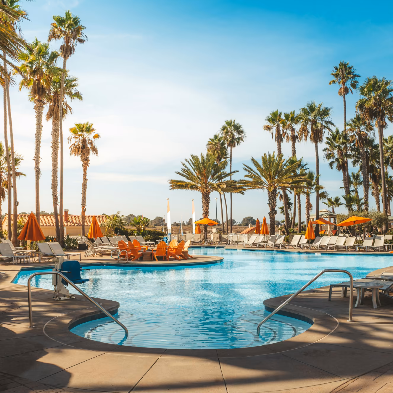 Resort swimming pool surrounded by palm trees, orange umbrellas, and lounge chairs under a clear blue sky.