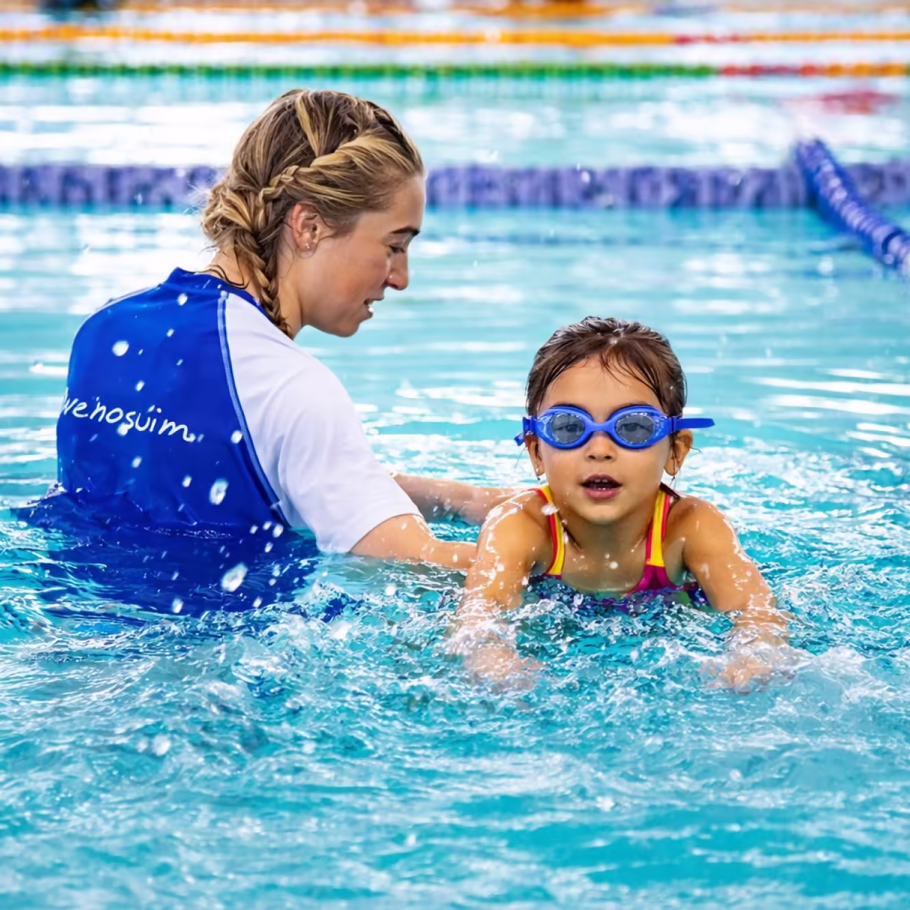 Swim instructor teaching a young girl wearing blue goggles to swim in a pool.