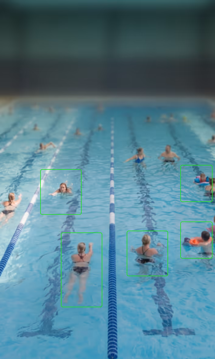 People of various ages enjoying swimming and playing in an indoor pool with onlookers seated nearby.