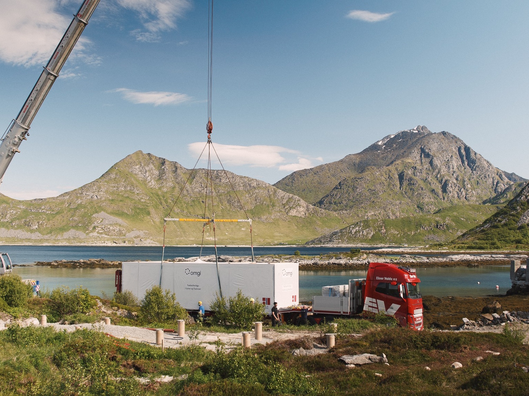 Kran løfter modulkonstruksjon fra lastebil ved vannet med fjell i bakgrunnen.