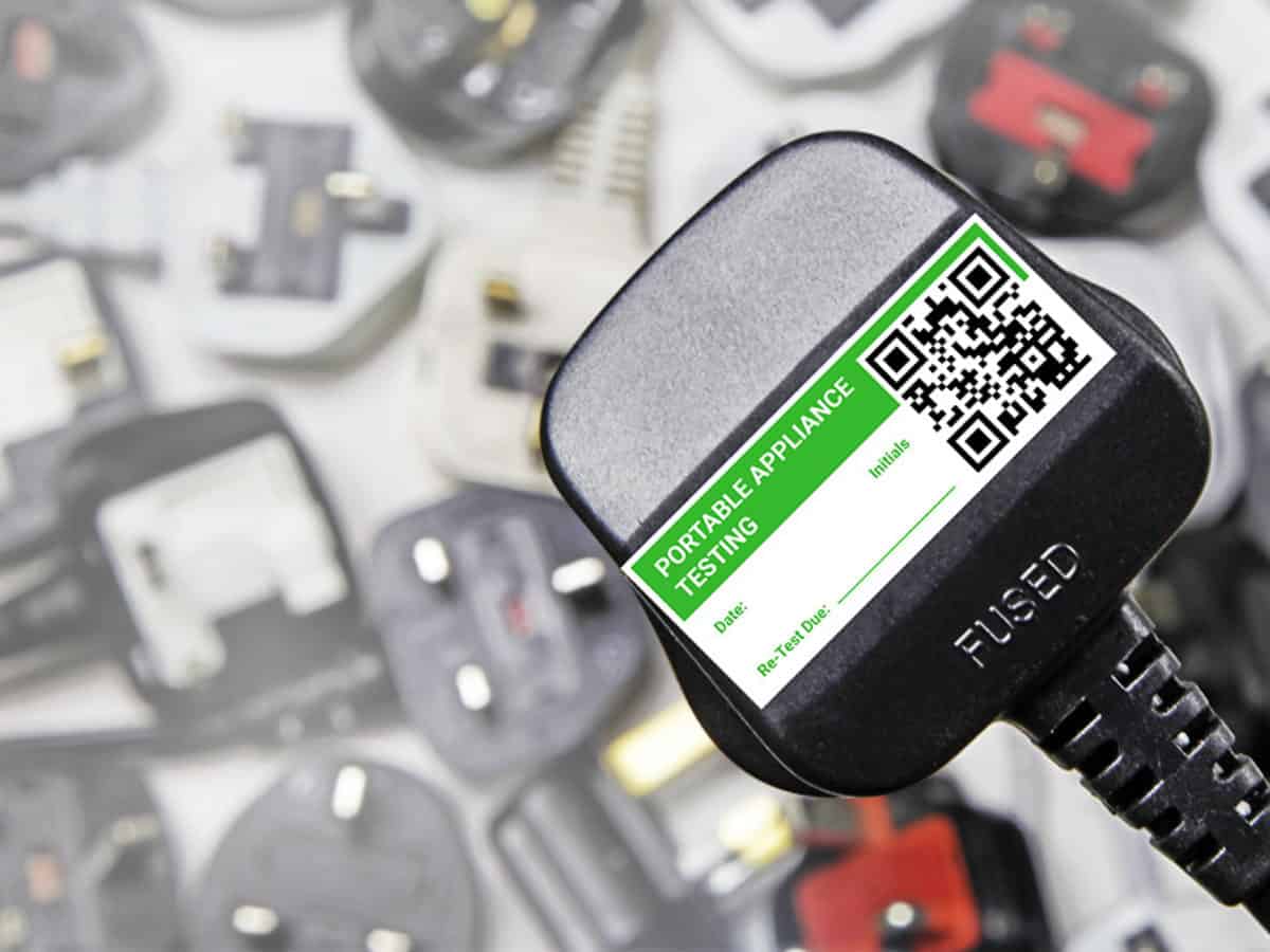 Black electrical plug with a green and white Portable Appliance Testing label and a QR code, surrounded by various blurred plugs in the background.