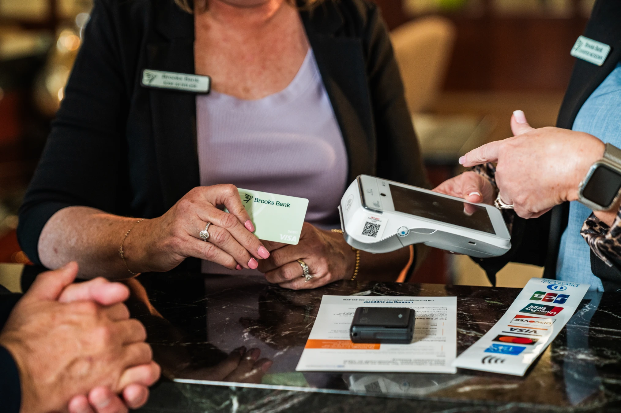 Brooks Bank banker handing a customer a debit card beside a card reader