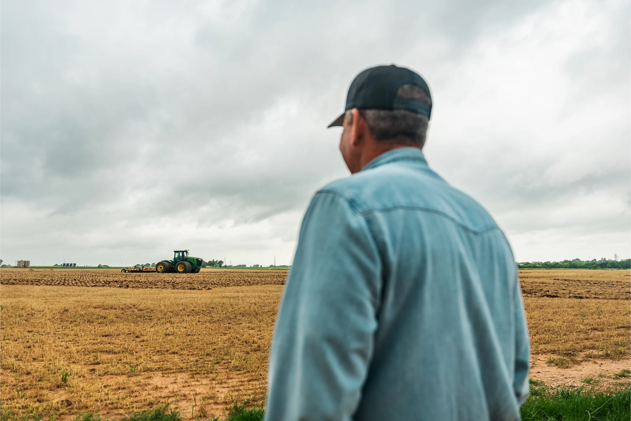 Farmer in a cap watching a tractor work a field under storm clouds