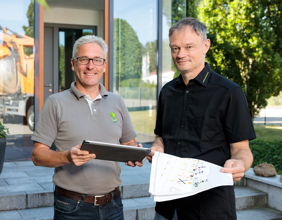 Zwei Männer stehen draußen vor einem Gebäude, einer hält ein Tablet, der andere einen Plan mit Markierungen.