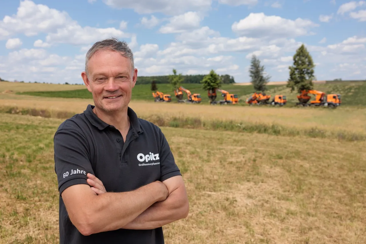 Mann mit schwarzen Poloshirt und verschränkten Armen steht auf einem Feld mit mehreren orangefarbenen Bau- oder Landmaschinen und Bäumen im Hintergrund.