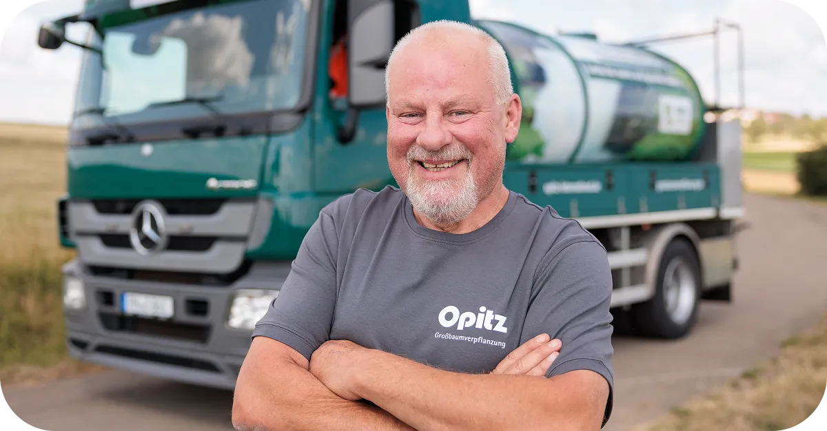 Älterer Mann mit grauem T-Shirt mit Opitz-Logo steht lächelnd mit verschränkten Armen vor einem grünen Mercedes-LKW.