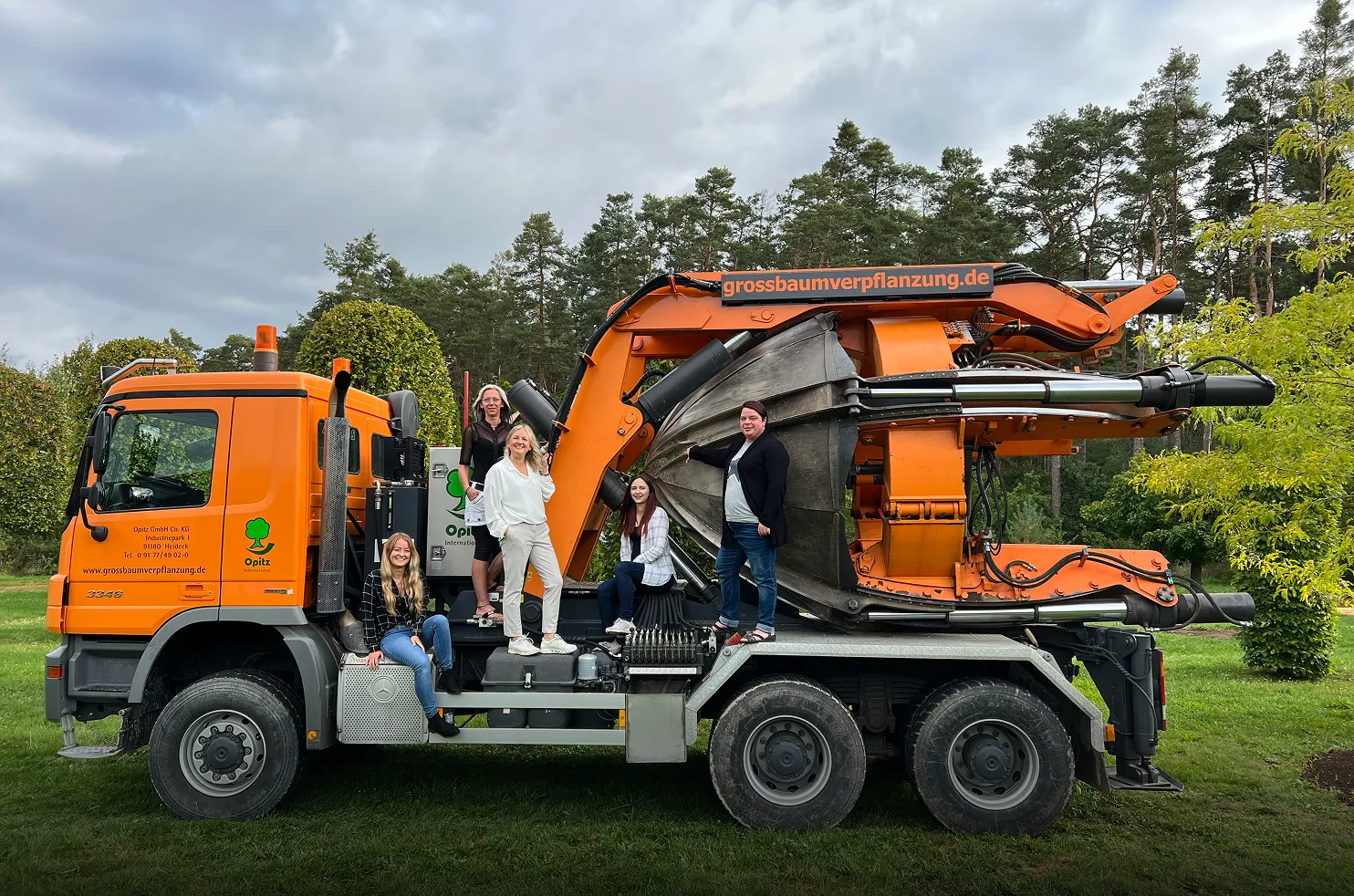 Fünf Frauen posieren auf einem orangen Baumtransporter mit großem Pflanzwerkzeug vor einem Wald.