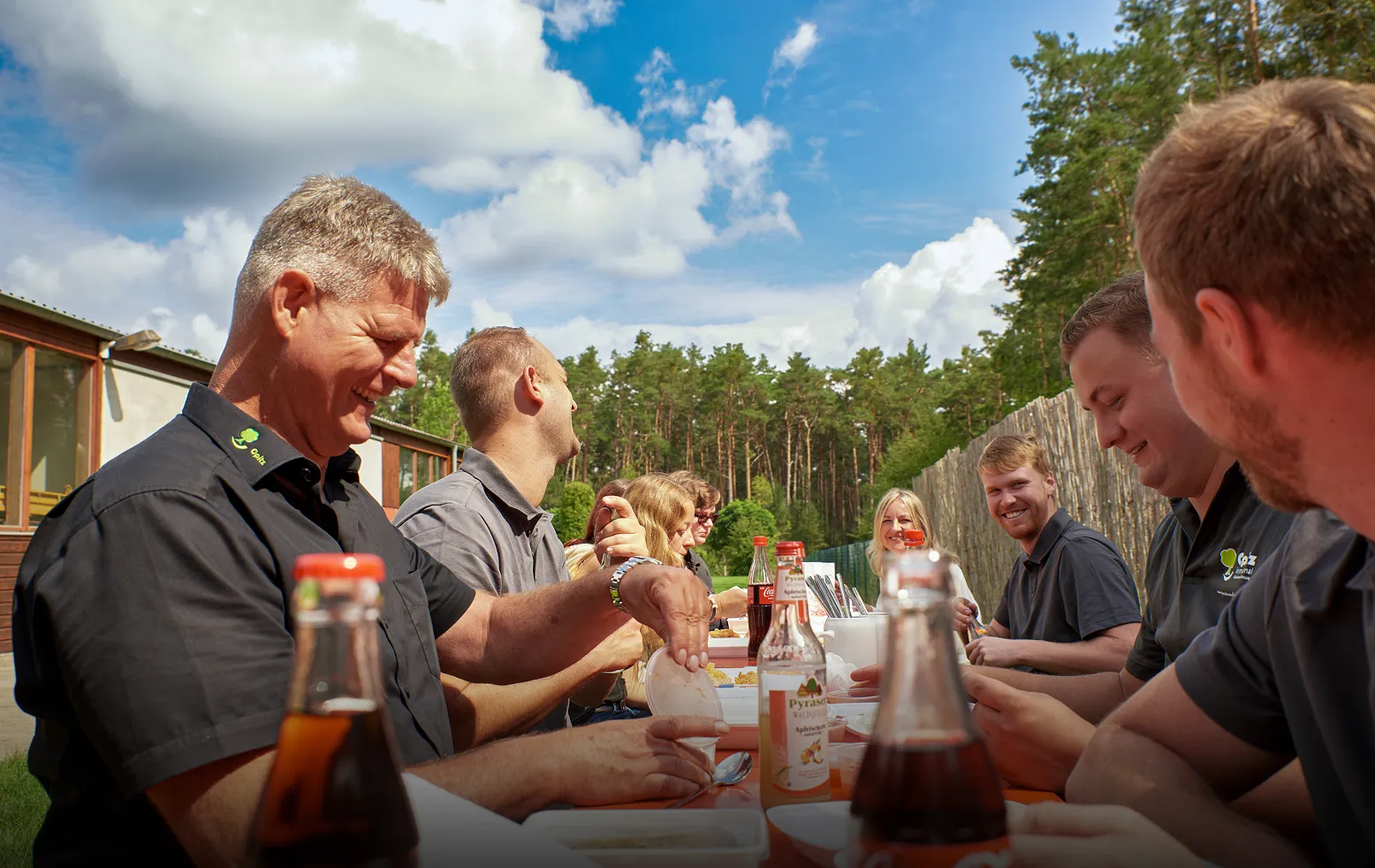 Gruppe von Menschen in Arbeitskleidung sitzt draußen an einem Tisch und isst gemeinsam bei sonnigem Wetter.