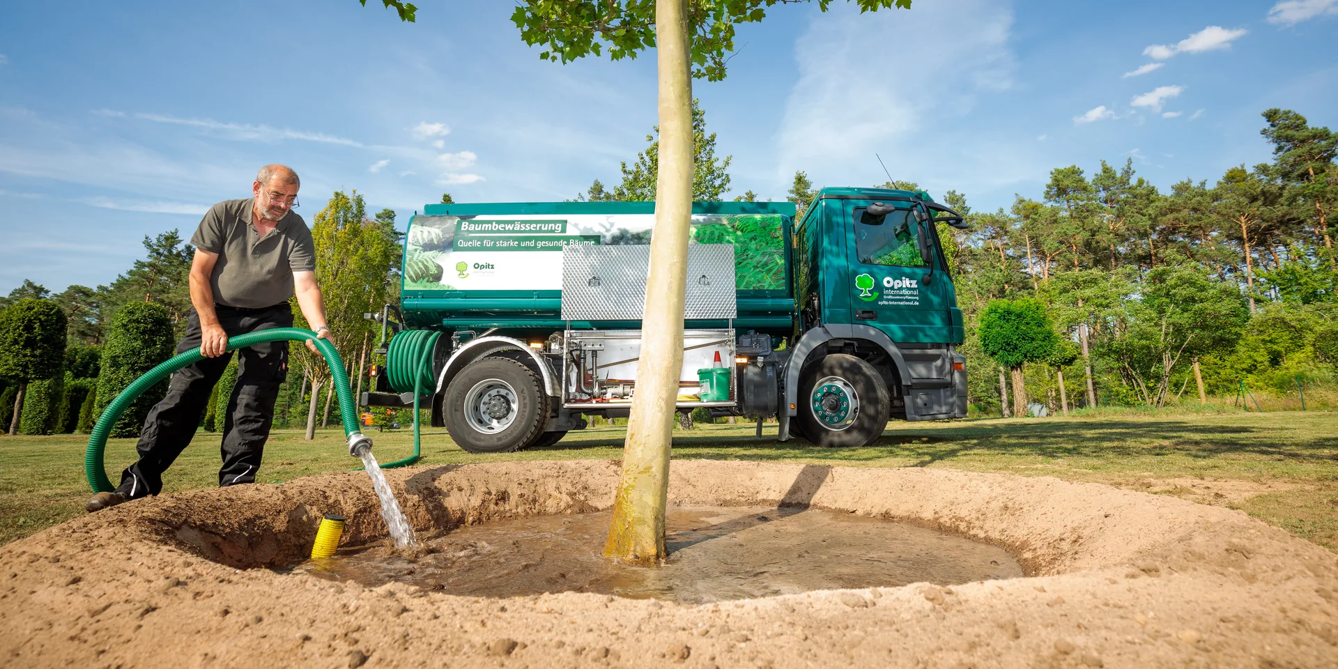 Mann bewässert einen Baum mit einem Schlauch, der an einem Wasserfahrzeug mit der Aufschrift Baumbewässerung befestigt ist.