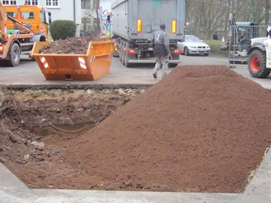 Großer aufgeschütteter Erdhaufen neben einem ausgehobenen Loch auf einer Baustelle mit Baufahrzeugen und einem Mann im Hintergrund.