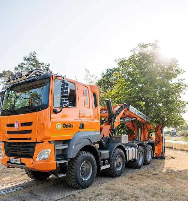 Oranger Baumverpflanzungslaster mit Kranarm steht bei sonnigem Wetter neben einem Baum.