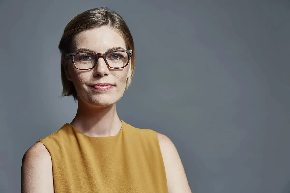 Confident woman wearing glasses and a mustard sleeveless top against a plain gray background.
