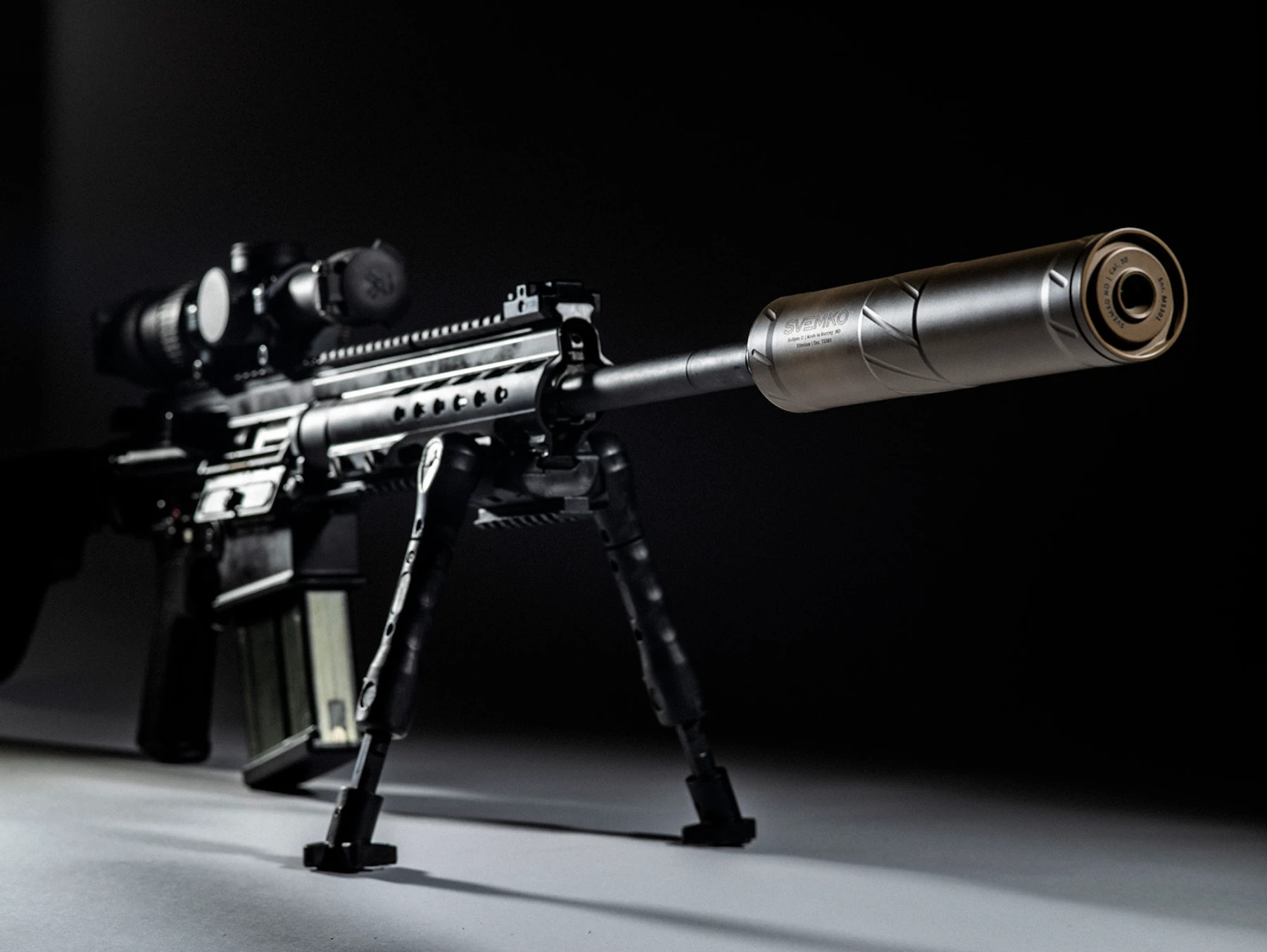 Tactical rifle with scope, bipod, and attached suppressor against black background.