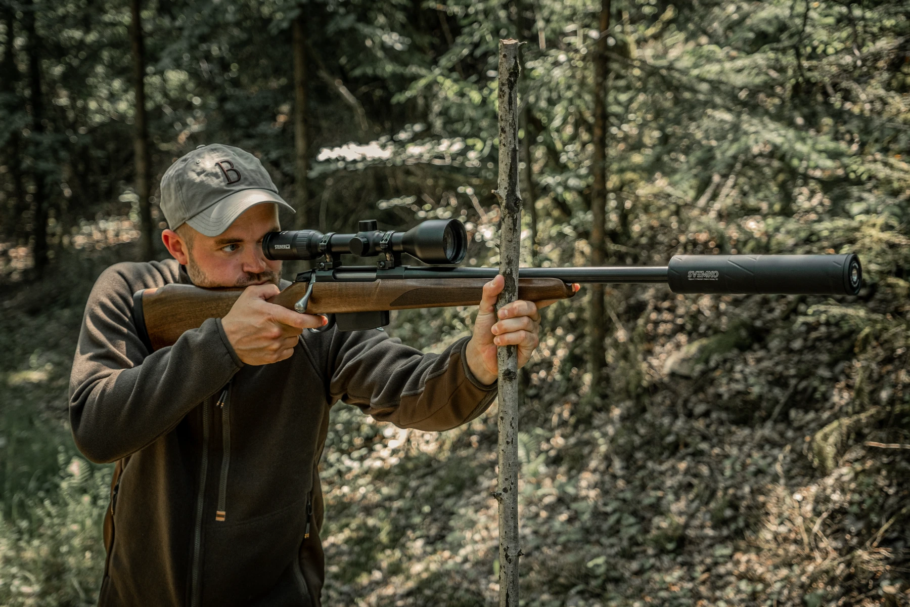 Man aiming a rifle with a scope and suppressor, using a tree branch for steadying, in a forest.