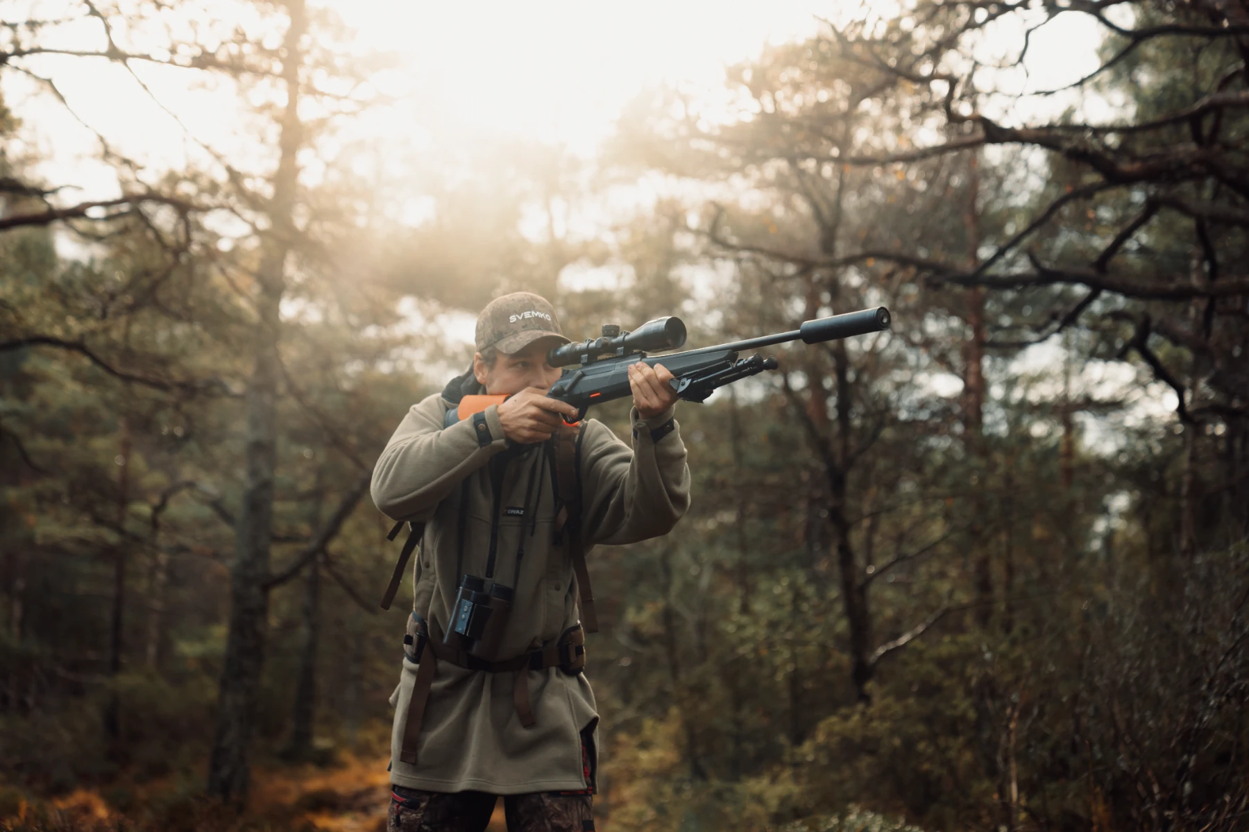Hunter aiming a scoped rifle with a suppressor in a forest at sunrise or sunset.
