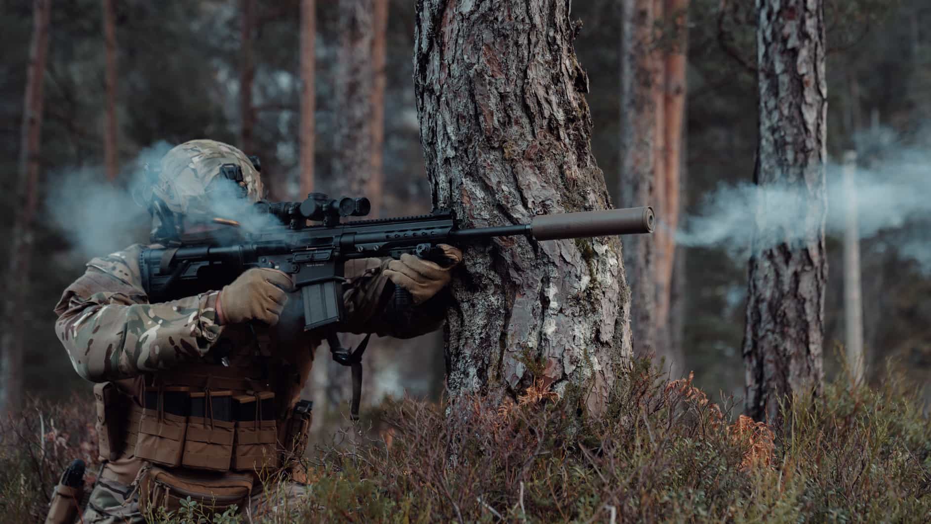 Soldier in camouflage gear firing a suppressed rifle in a forest, smoke visible around the barrel.