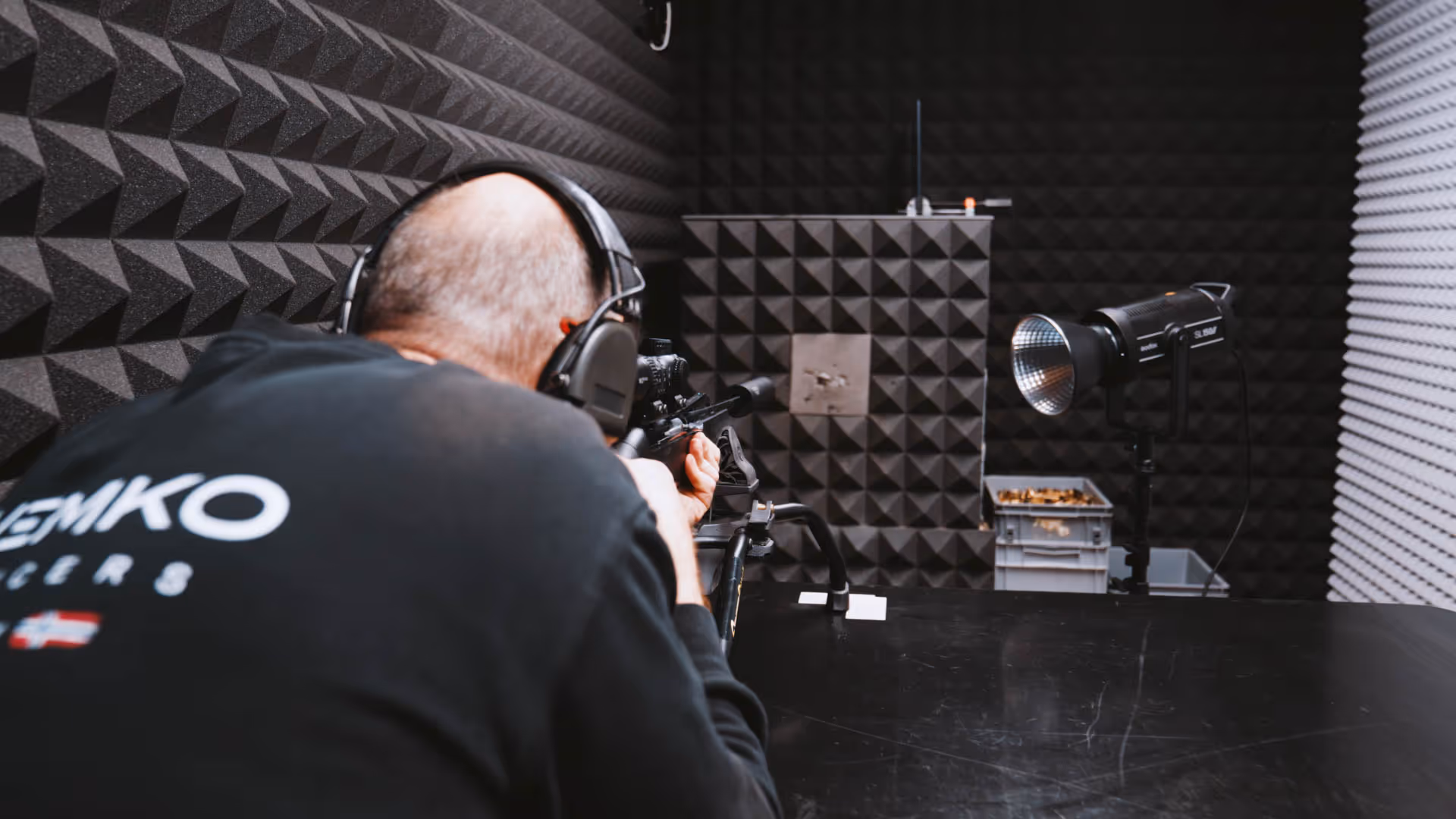 Man wearing headphones aiming a rifle at a bulletproof target in a soundproofed indoor shooting range.