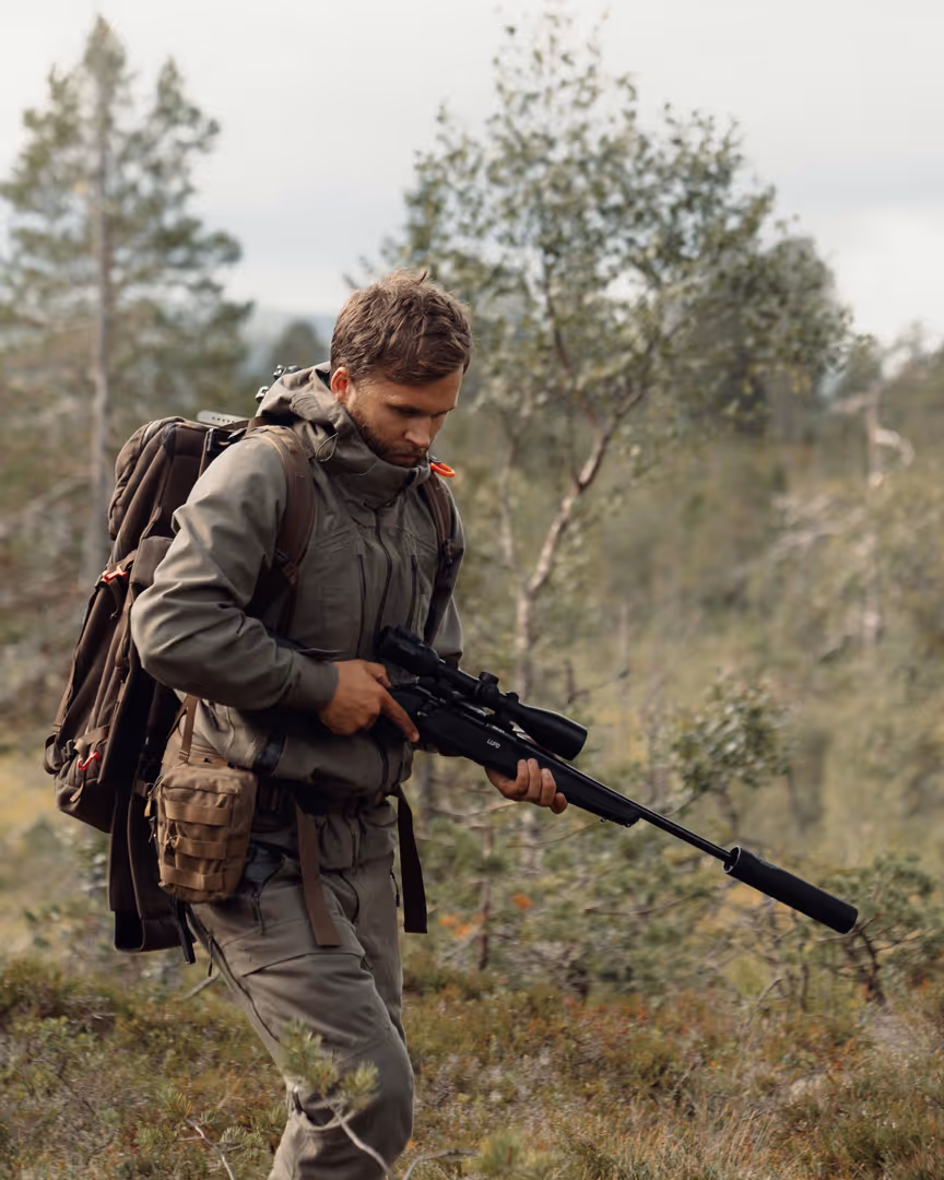 Man wearing outdoor gear and backpack holding a scoped rifle with silencer in a forest.