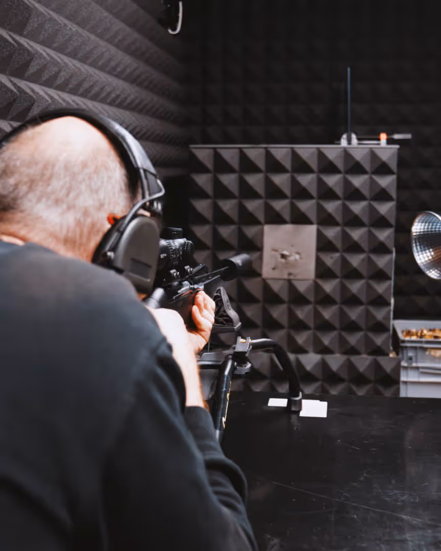 Person wearing black headphones aiming a scoped rifle at a target in a soundproof shooting range.
