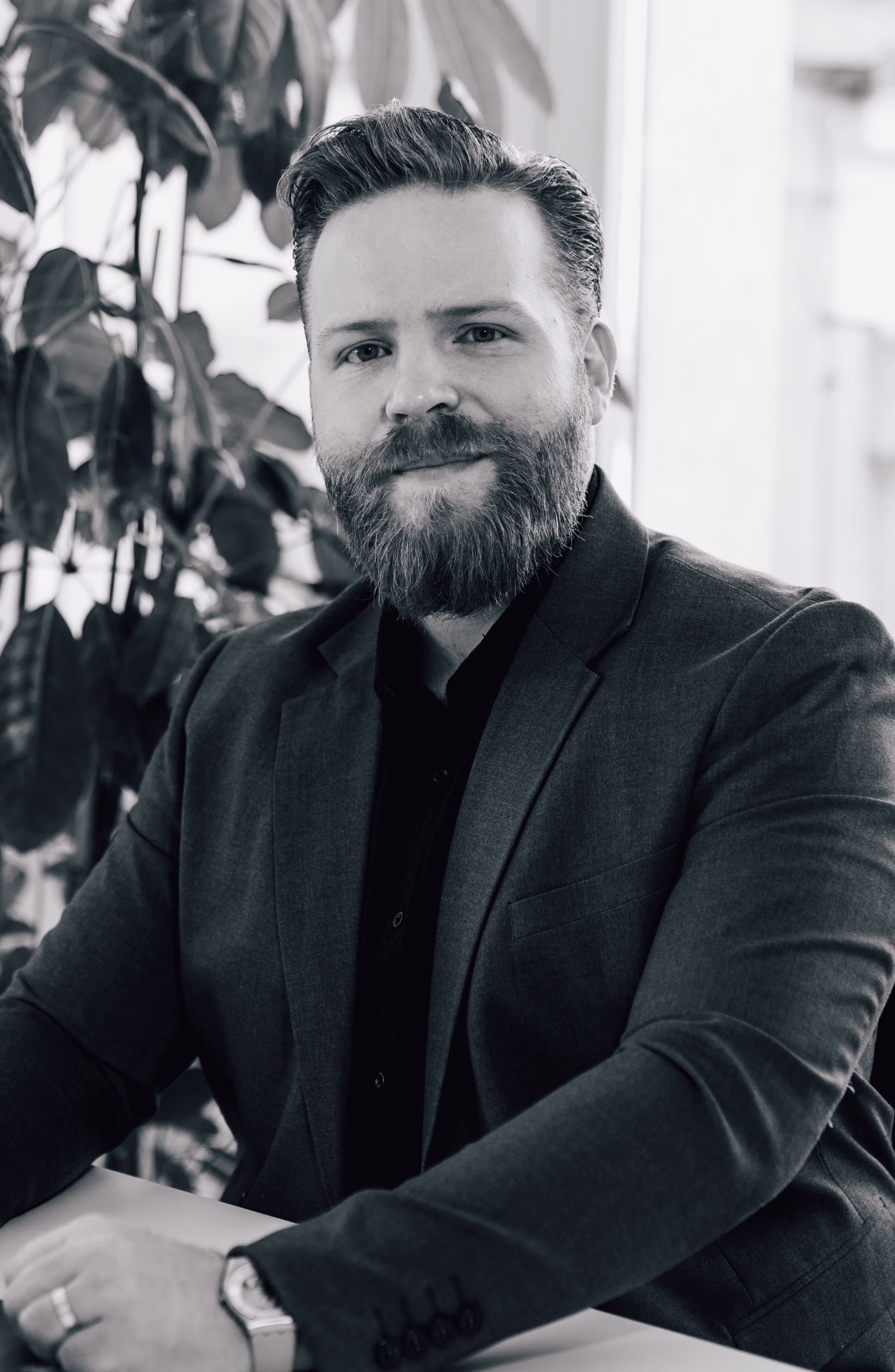 Porträt eines bärtigen Mannes in Anzug, der entspannt an einem Tisch sitzt mit Pflanzen im Hintergrund.