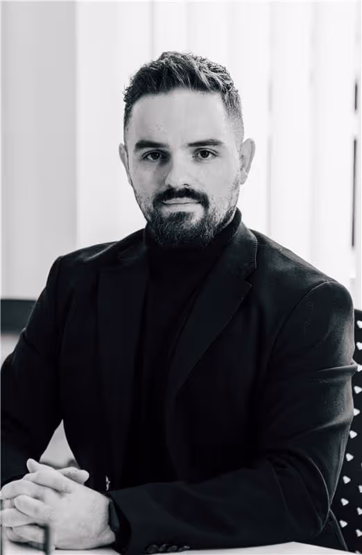 Porträt eines Mannes mit dunklem Haar und Bart, der einen schwarzen Anzug und Rollkragenpullover trägt, sitzt an einem Tisch mit verschränkten Händen.