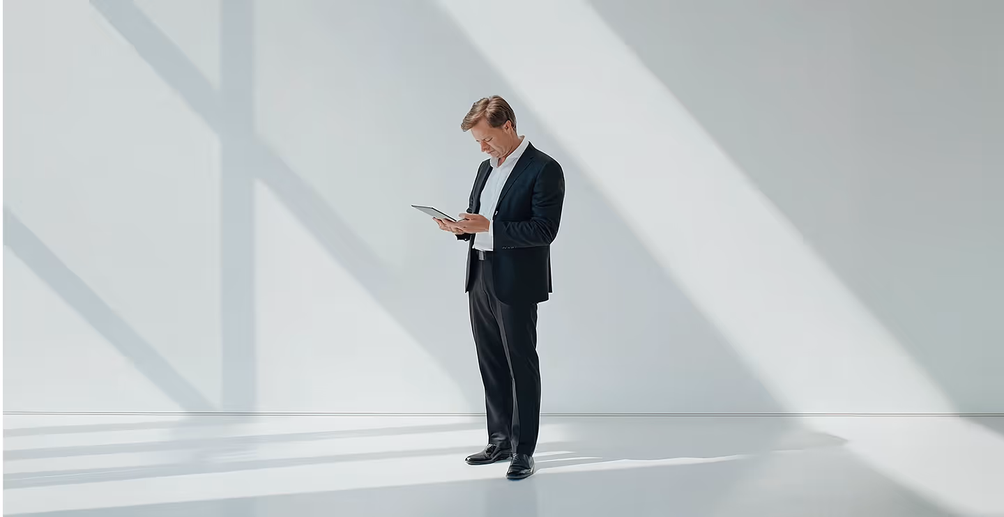 Geschäftsmann in schwarzem Anzug steht in hellem Raum und schaut auf ein Tablet.