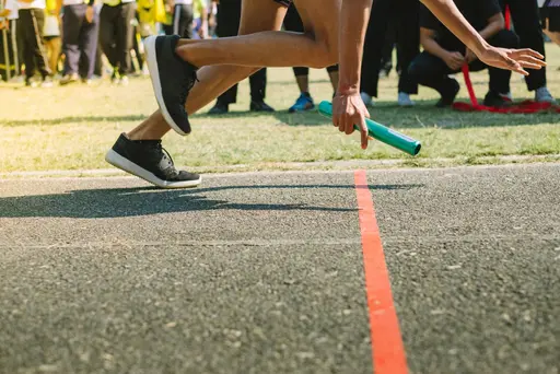 Athlete holding a relay baton and sprinting on a track near a red line, with spectators in the background.