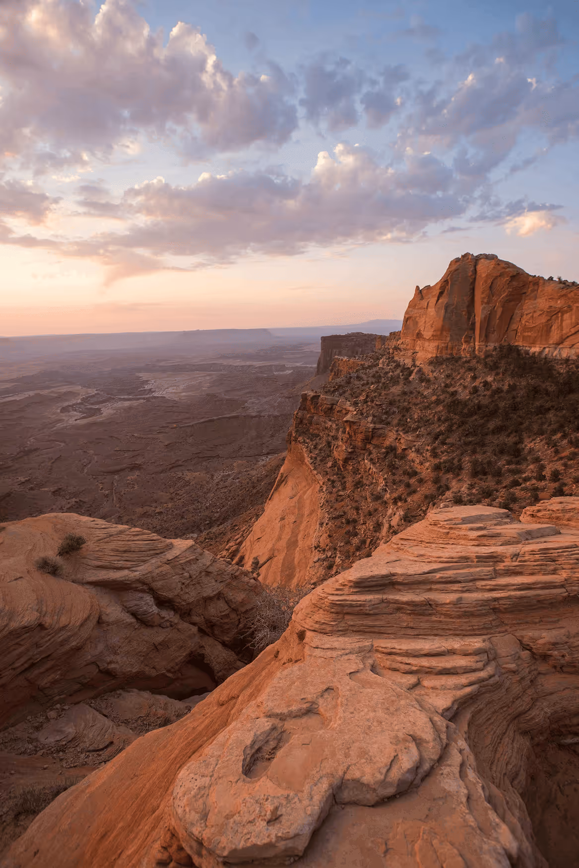 Sunset over a rocky desert canyon with layered red sandstone cliffs and sparse vegetation under a partly cloudy sky.