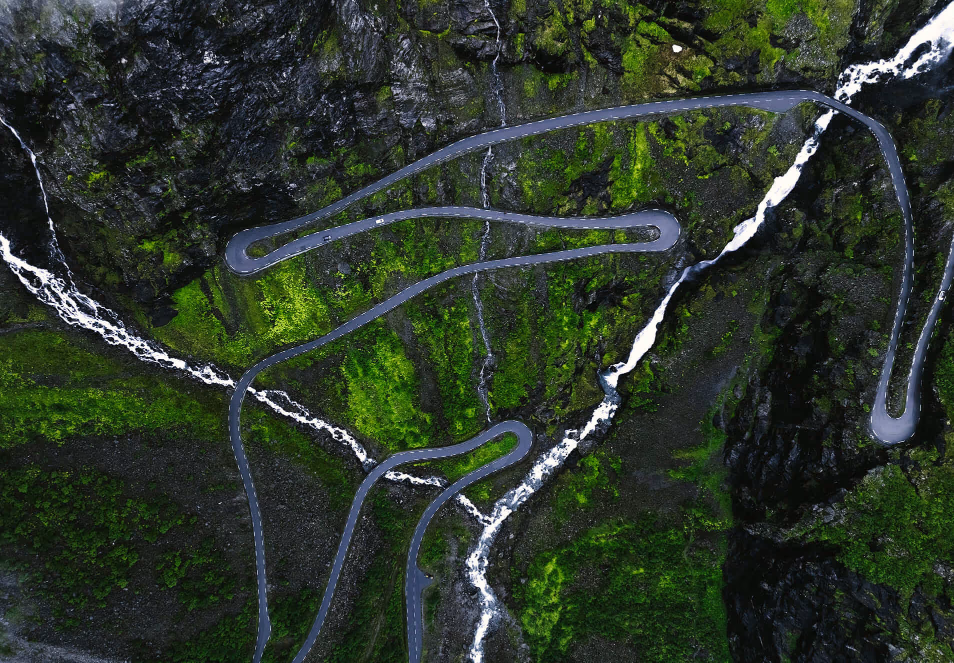 Aerial view of a winding mountain road surrounded by green moss-covered rocks and multiple cascading waterfalls.