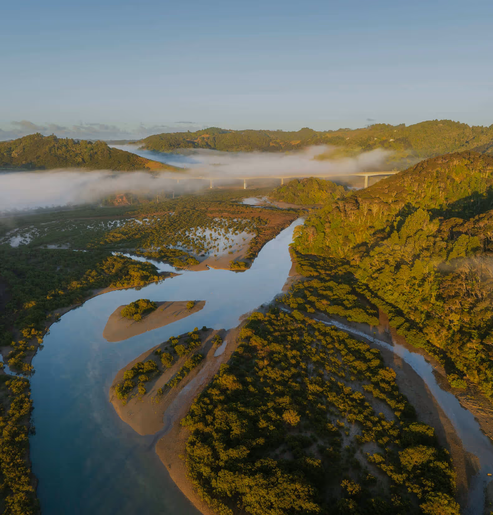 A winding river flowing through dense green forested hills with mist and a bridge in the background under a clear sky.