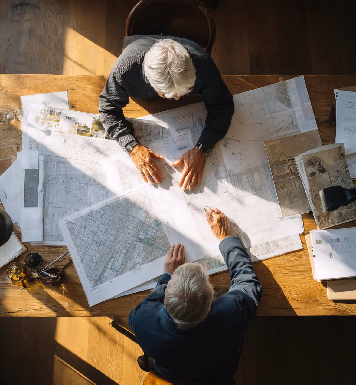 Two elderly men with white hair sitting at a wooden table reviewing large architectural blueprints and maps under sunlight.