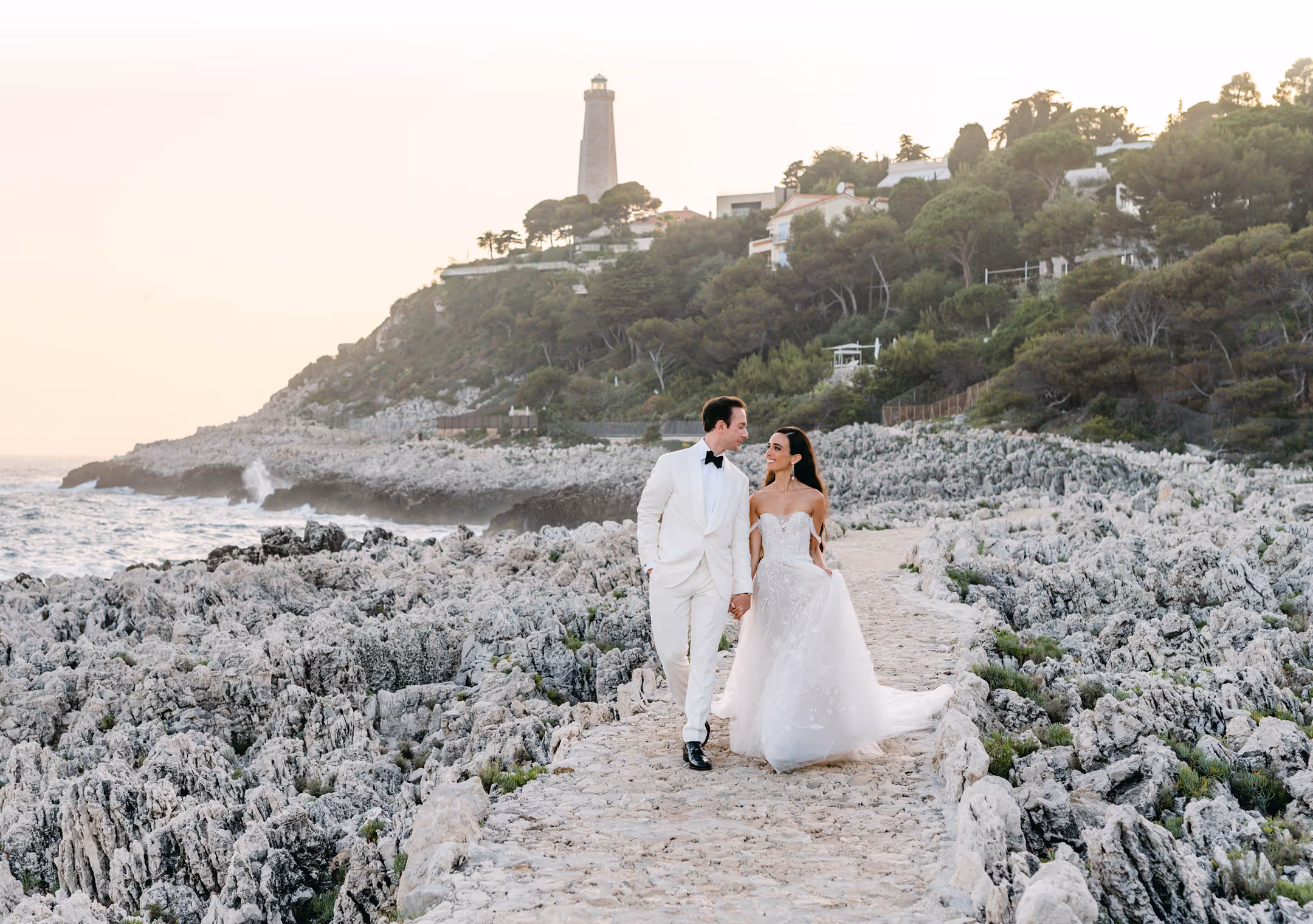 Bride and groom holding hands and walking on a rocky seaside path with a lighthouse and trees on a hill in the background.