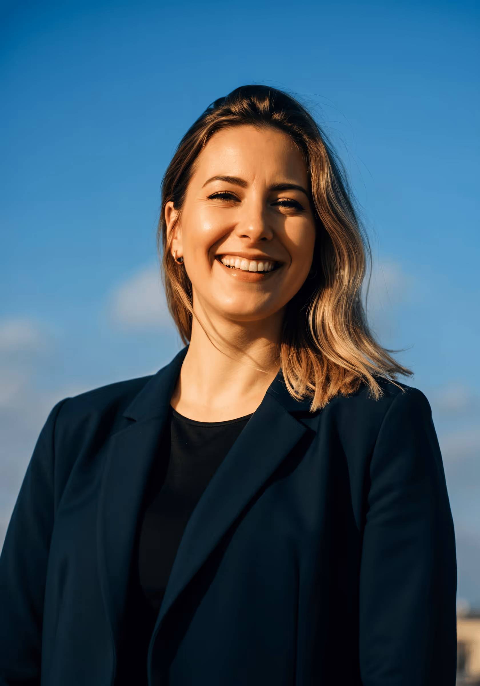 Smiling woman with shoulder-length blonde hair wearing a dark blazer against a clear blue sky.