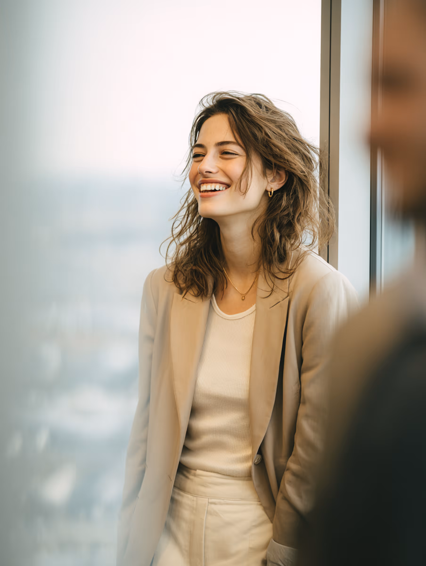 Young woman with wavy hair smiling and standing by a window wearing a beige blazer and white outfit.