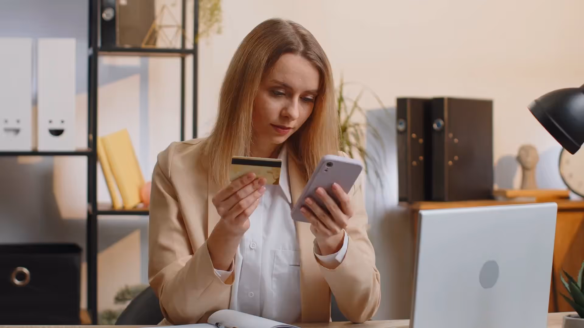 Woman holding a credit card and smartphone, sitting at a desk with a laptop in a home office.