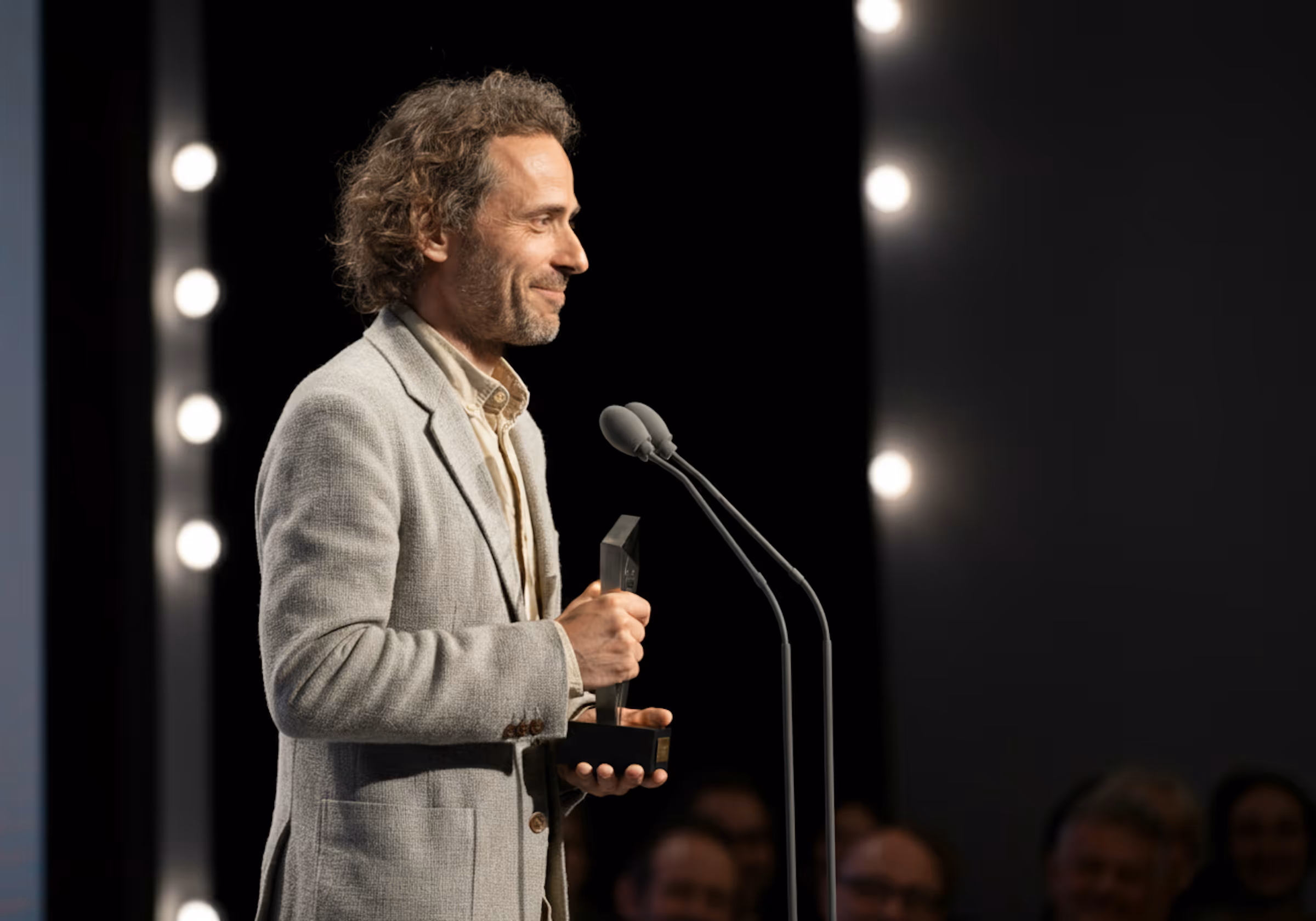 Man in light gray blazer holding an award and speaking at a microphone on stage.