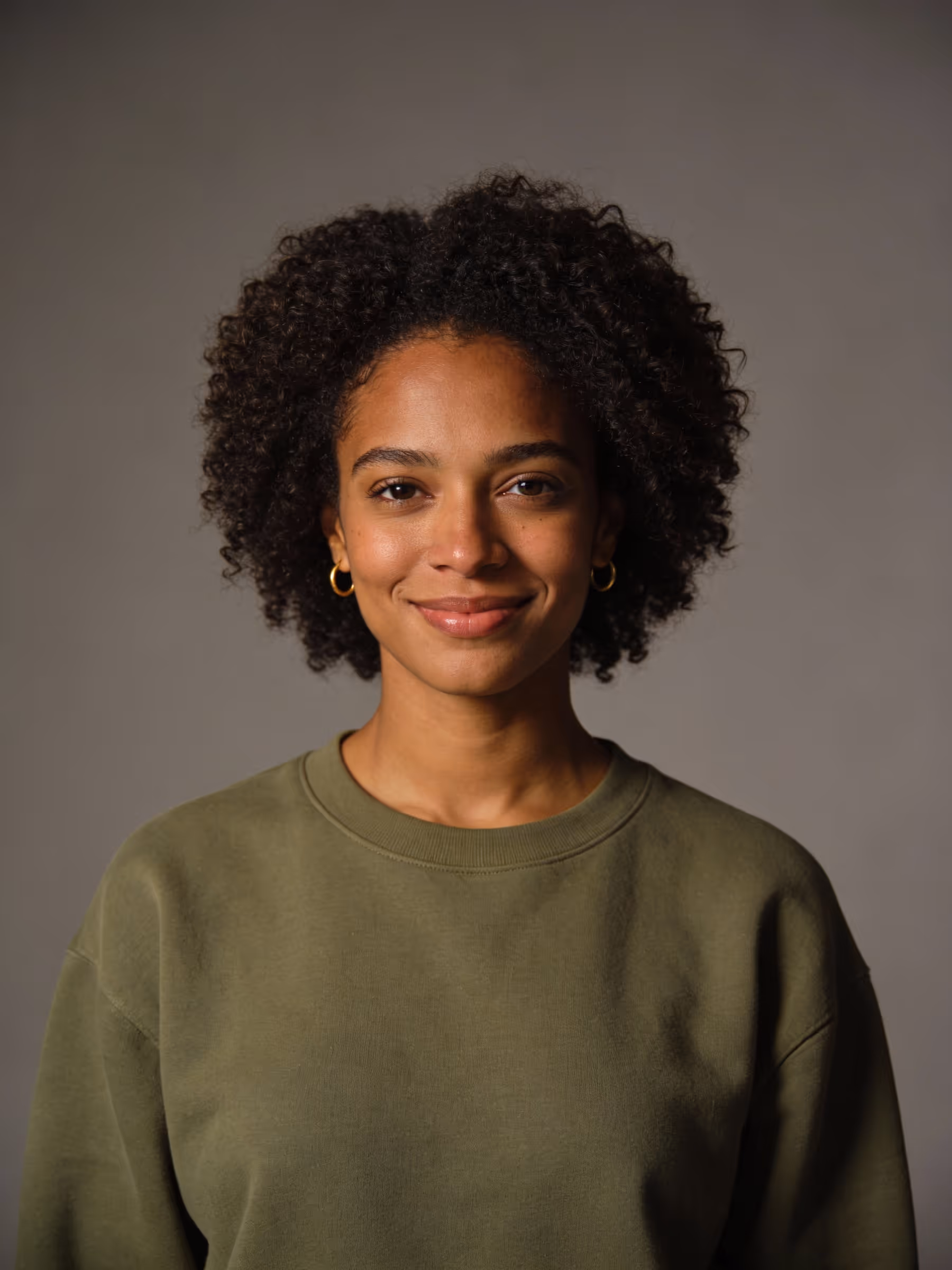 Smiling woman with curly hair wearing a green sweatshirt and gold hoop earrings against a plain background.