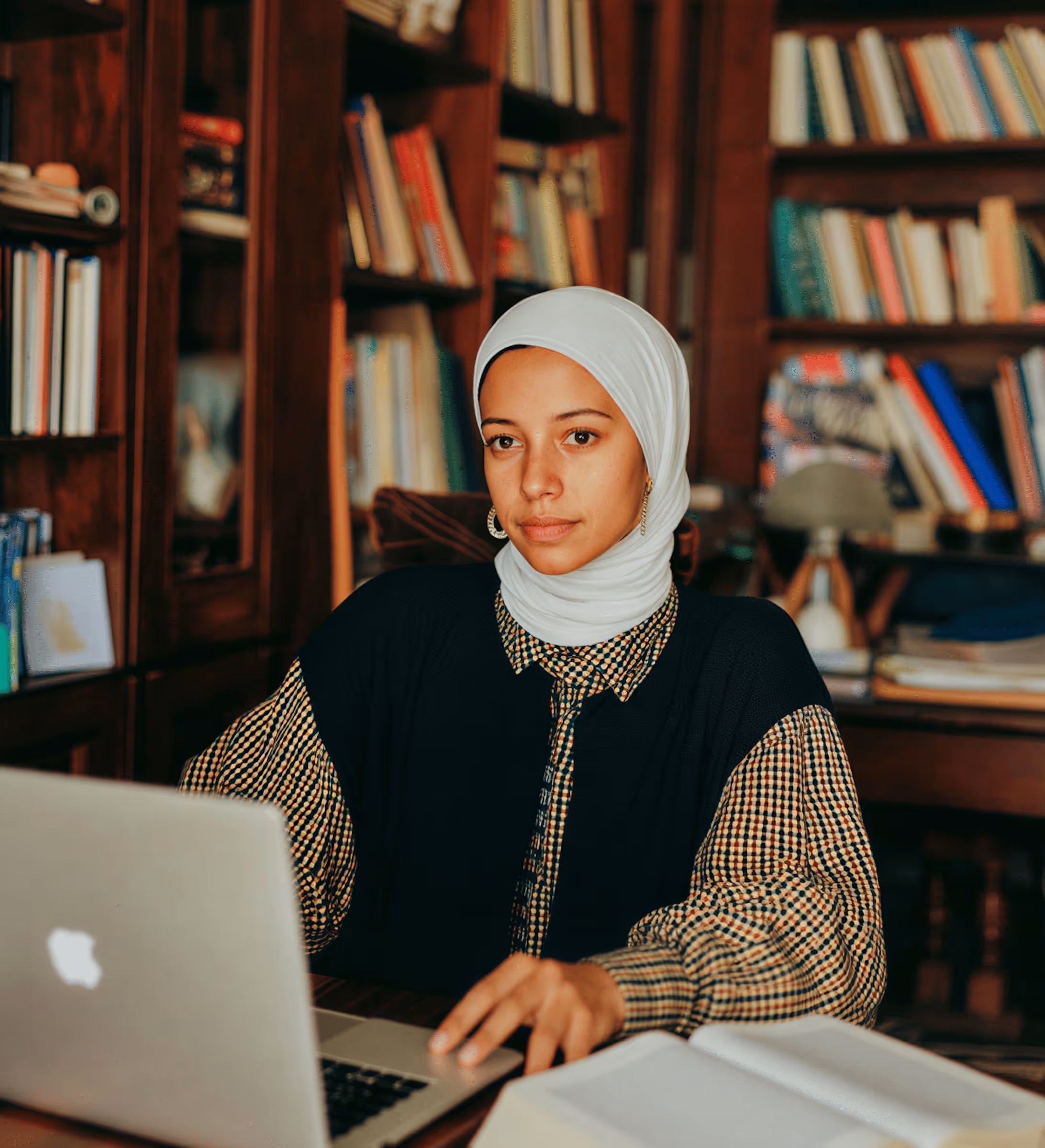 Woman wearing white hijab working on a laptop in a library with bookshelves behind her.
